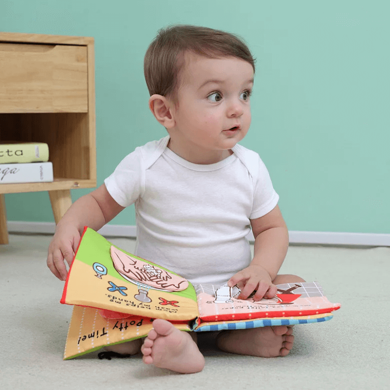 Little boy sitting on a carpet and holding a colorful potty training- themed soft book. 